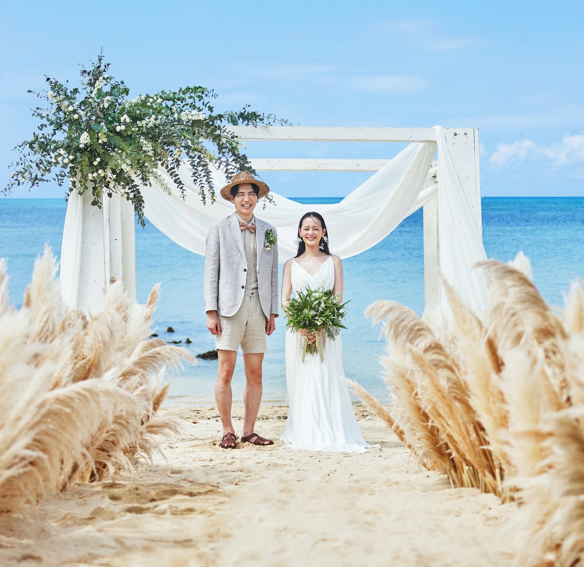 パンパスグラスが飾られた流木のアートの前で、青い海を背に微笑む新郎新婦。麦わら帽子にハーフパンツスタイルの新郎と、ナチュラルな白いドレスに花びらのようなイヤリングを合わせた新婦が、沖縄の自然を感じる砂浜で佇む美しいビーチフォトです。