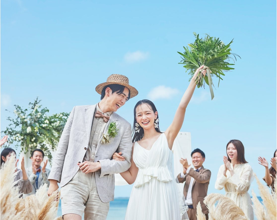 青い海を背景にしたビーチウエディングで、幸せそうな新郎新婦と、二人を笑顔で見守る参列者たち