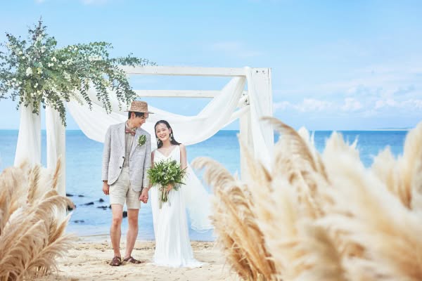 エメラルドグリーンの海が広がるビーチの祭壇で、カメラに笑顔を向ける新婦と、その隣で優しく見つめる新郎。パンパスグラスが揺れる砂浜のバージンロードで、麦わら帽子を被ったカジュアルな新郎と、グリーンのブーケを持つ新婦が幸せな時を刻むビーチウエディングの一枚です。