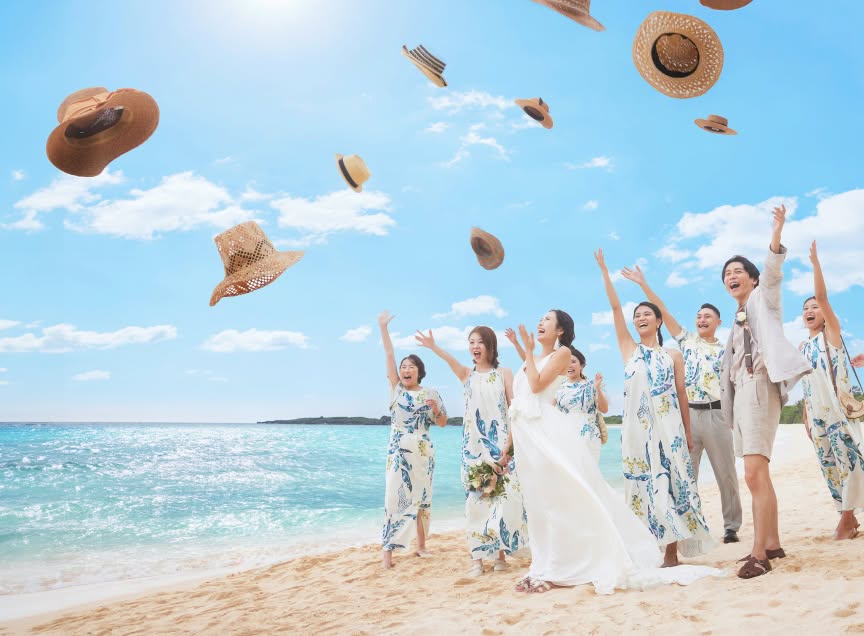 澄み渡る青い空に向かって、新郎新婦とゲスト全員が麦わら帽子を一斉に投げ上げる歓喜の瞬間。白い砂浜とエメラルドグリーンの海が広がる絶好のロケーションで、お揃いのかりゆしワンピースを着た友人たちと楽しむ、開放感あふれる沖縄のビーチウエディングのワンシーンです。