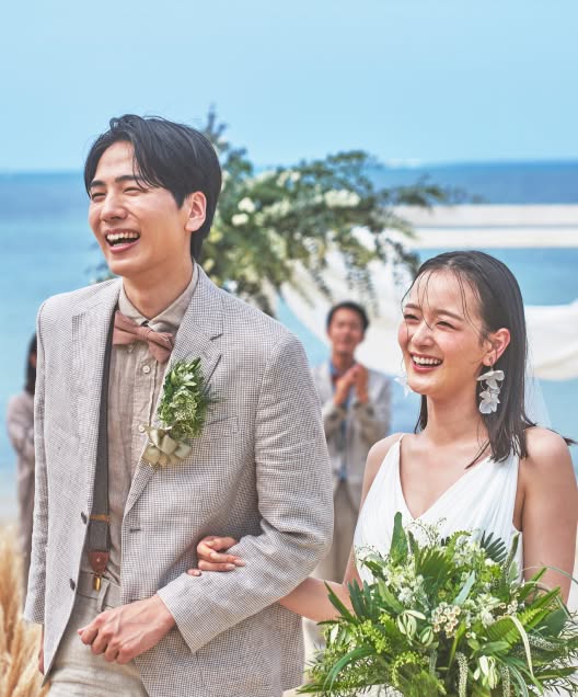 エメラルドグリーンの海を背景にしたビーチの祭壇で、ゲストの祝福を受けながら弾けるような笑顔を見せる新郎新婦。麦わら帽子を被ったカジュアルなスタイルの新郎と、大ぶりのフラワーイヤリングが揺れる白ドレス姿の新婦が、開放感あふれる沖縄の空の下で幸せを噛みしめる瞬間です。