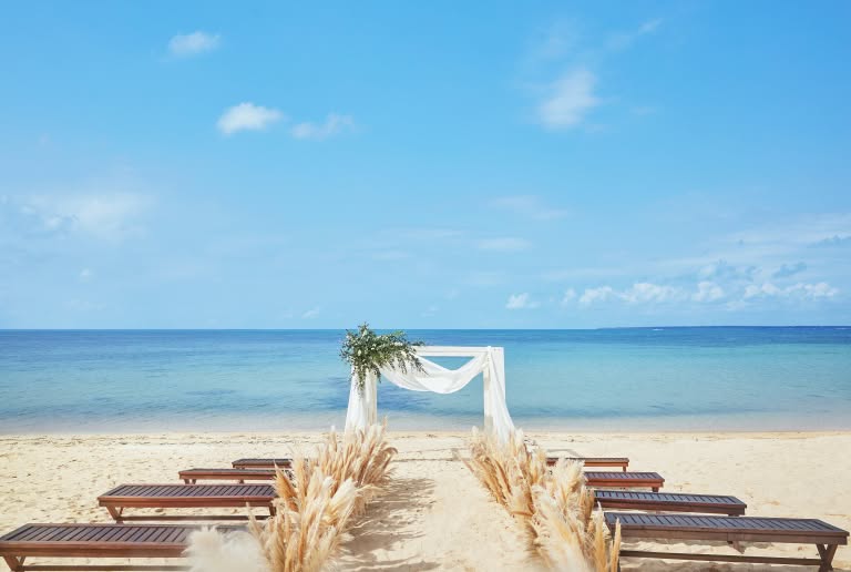 透き通るエメラルドグリーンの海を背景に、砂浜に設営された開放的なビーチウエディング会場。流木とグリーンで装飾された白い木製の祭壇と、バージンロード沿いに並ぶ木製のベンチが、ナチュラルで上質なリゾートセレモニーを演出する全景カットです。