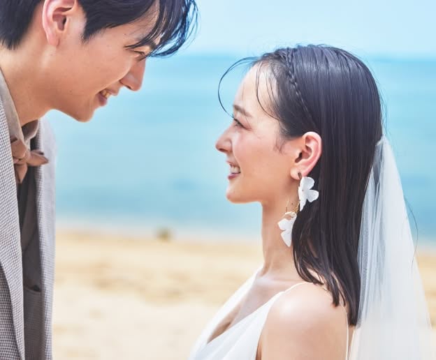 穏やかな沖縄の海を背景に、至近距離で見つめ合い幸せそうに微笑む新郎新婦。白い花びらのような大ぶりのイヤリングをつけた新婦と、優しい表情の新郎が、ビーチでのロケーションフォトで愛おしそうに視線を交わす心温まるクローズアップショットです。
