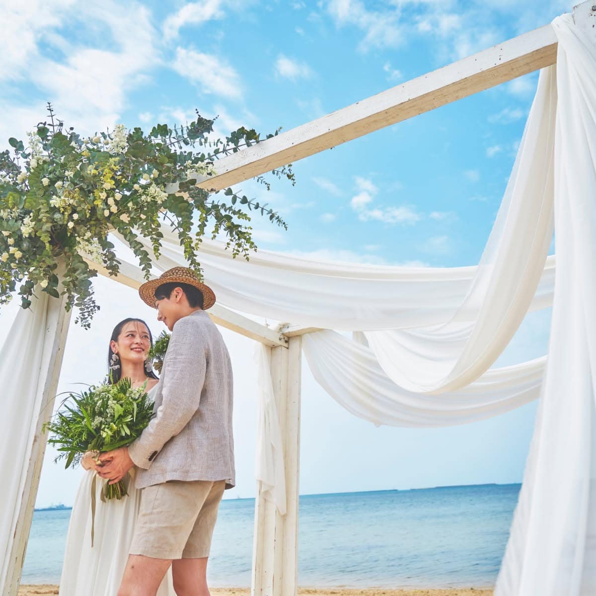 青空に映える白い布とグリーンで装飾されたビーチの祭壇で、寄り添いながら見つめ合う新郎新婦。麦わら帽子を被ったカジュアルな新郎と、ナチュラルなドレスを纏った新婦が、沖縄の開放的な海を背景に穏やかな時間を過ごしている。