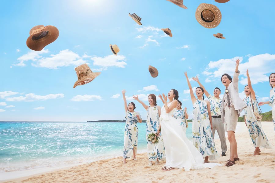 澄み渡る青い空に向かって、新郎新婦とゲスト全員が麦わら帽子を一斉に投げ上げる歓喜の瞬間。白い砂浜とエメラルドグリーンの海が広がる絶好のロケーションで、お揃いのかりゆしワンピースを着た友人たちと楽しむ、開放感あふれる沖縄のビーチウエディングのワンシーンです。