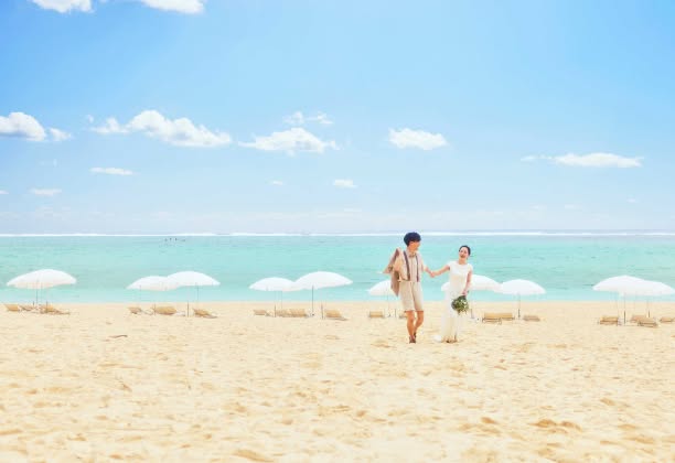 果てしなく広がる白い砂浜とエメラルドグリーンの海を背景に、点在する白いパラソルの間を仲睦まじく歩く新郎新婦。沖縄の自然美をパノラマで捉えた、開放感あふれるビーチロケーションフォトの全景シーンです。