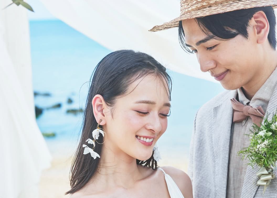 ビーチに設置された白い布の祭壇の下で、目を伏せて微笑む新婦を、新郎が隣で愛おしそうに見つめるクローズアップショット。麦わら帽子を被った新郎と、大ぶりの白い花びらイヤリングをつけた新婦が、沖縄の穏やかな海を背景に幸せを噛みしめる瞬間です。