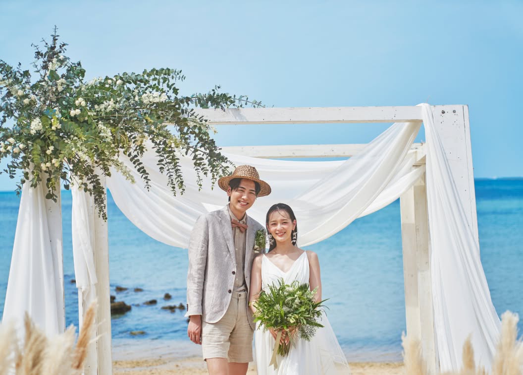 エメラルドグリーンの海を背景にした白い木製の祭壇で、寄り添って正面を見つめる新郎新婦。麦わら帽子を被ったナチュラルなスタイルの新郎と、ボリュームのあるグリーンのブーケを抱えた新婦が、沖縄の柔らかな光に包まれるビーチウエディングのポートレートです。