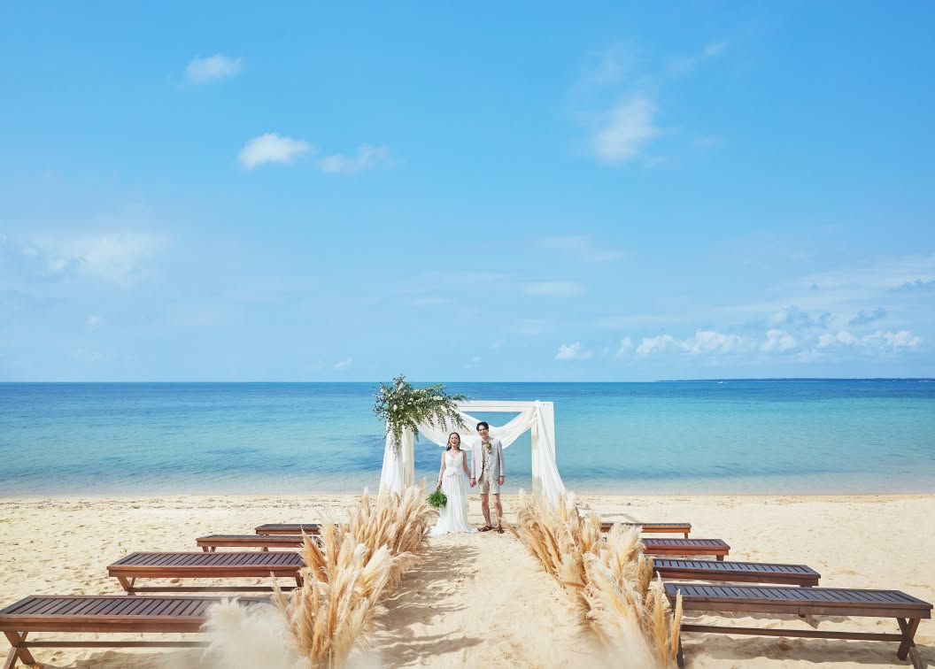 どこまでも続く青い海と空を背景に、砂浜に設置された流木とパンパスグラスの祭壇。木製のベンチが並び、自然の素材を活かした装飾が美しい、沖縄のロケーションを贅沢に活用した開放感あふれるビーチウエディングの会場全景です。