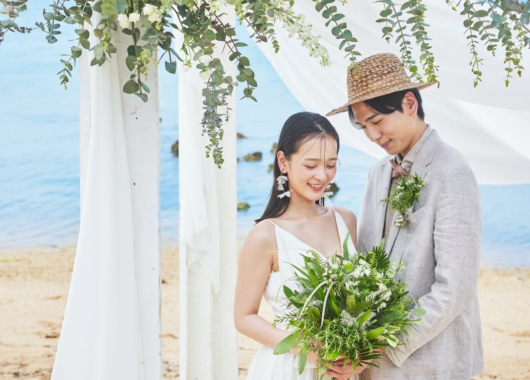 沖縄の柔らかな光の中、ウェディングベールをなびかせて新郎と見つめ合う新婦。大ぶりの白いフラワーイヤリングが印象的な新婦と、穏やかな眼差しを向ける新郎の横顔を捉えた、ビーチでの親密な空気感が伝わるクローズアップフォトです。