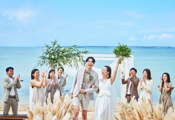 沖縄の青い海を背に、ゲストの拍手に包まれて笑顔で退場する新郎新婦。右手にグリーンのブーケを高く掲げる新婦と、腕を組みながら晴れやかな表情を浮かべる新郎が、開放感あふれるビーチで祝福を浴びる幸せなフィナーレの瞬間です。