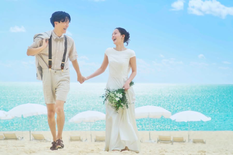 きらめくエメラルドグリーンの海を背に、砂浜で手をつないで楽しそうに歩く新郎新婦。サスペンダーにハーフパンツスタイルの新郎と、シンプルなフレンチスリーブの白ドレスを纏った新婦が、開放感あふれる沖縄のビーチで笑顔を交わす幸せなロケーションフォトです。