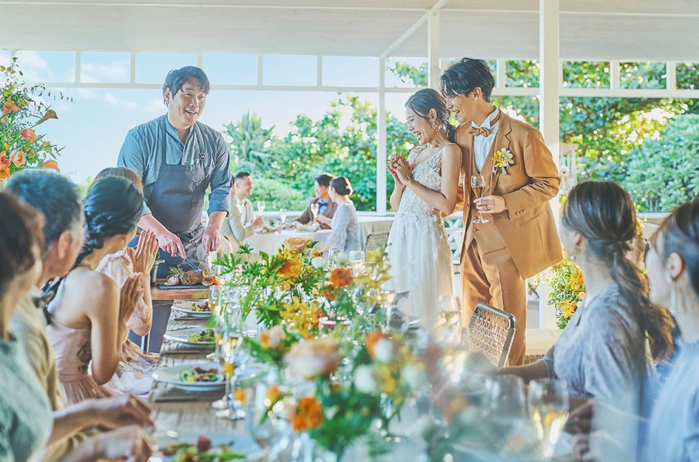 自然光が差し込む披露宴会場で、シェフが新郎新婦の目の前で肉料理を切り分けながら説明し、周囲のゲストが笑顔で拍手している様子