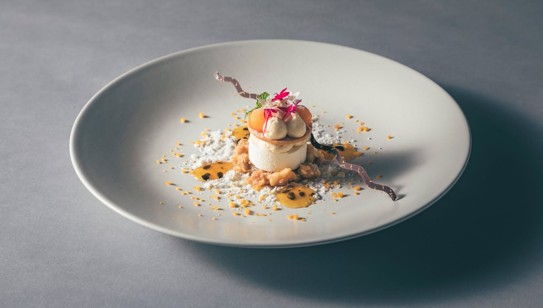 アイネス ヴィラノッツェ 沖縄のコースを締めくくる、パッションフルーツとココナッツのウエディングデザート。白いムースの上にマンゴーやエディブルフラワーが飾られ、波のようなチョコレートと南国らしいソースが彩る、華やかな一皿。