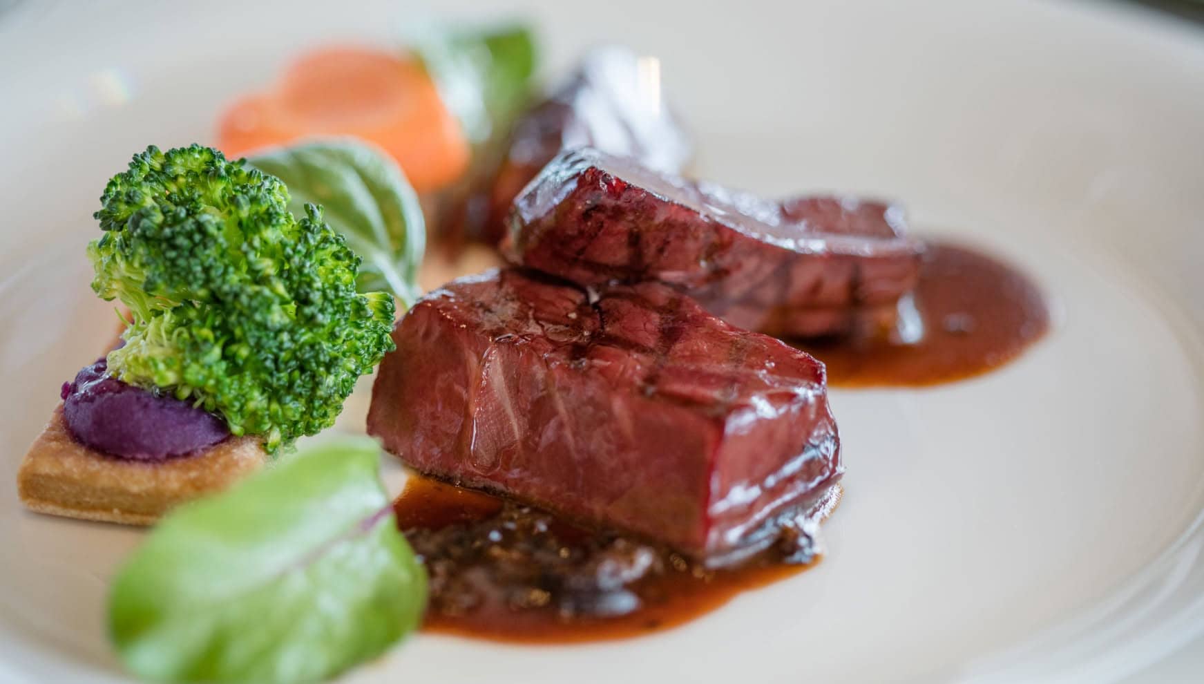 アイネス ヴィラノッツェ in カヌチャベイで提供される、牛フィレ肉のグリル。絶妙な焼き加減の肉料理に、ブロッコリーや紫芋のピューレ、彩り豊かな野菜を添え、濃厚なソースが味わいを引き立てる、華やかなウエディングのメインディッシュ。