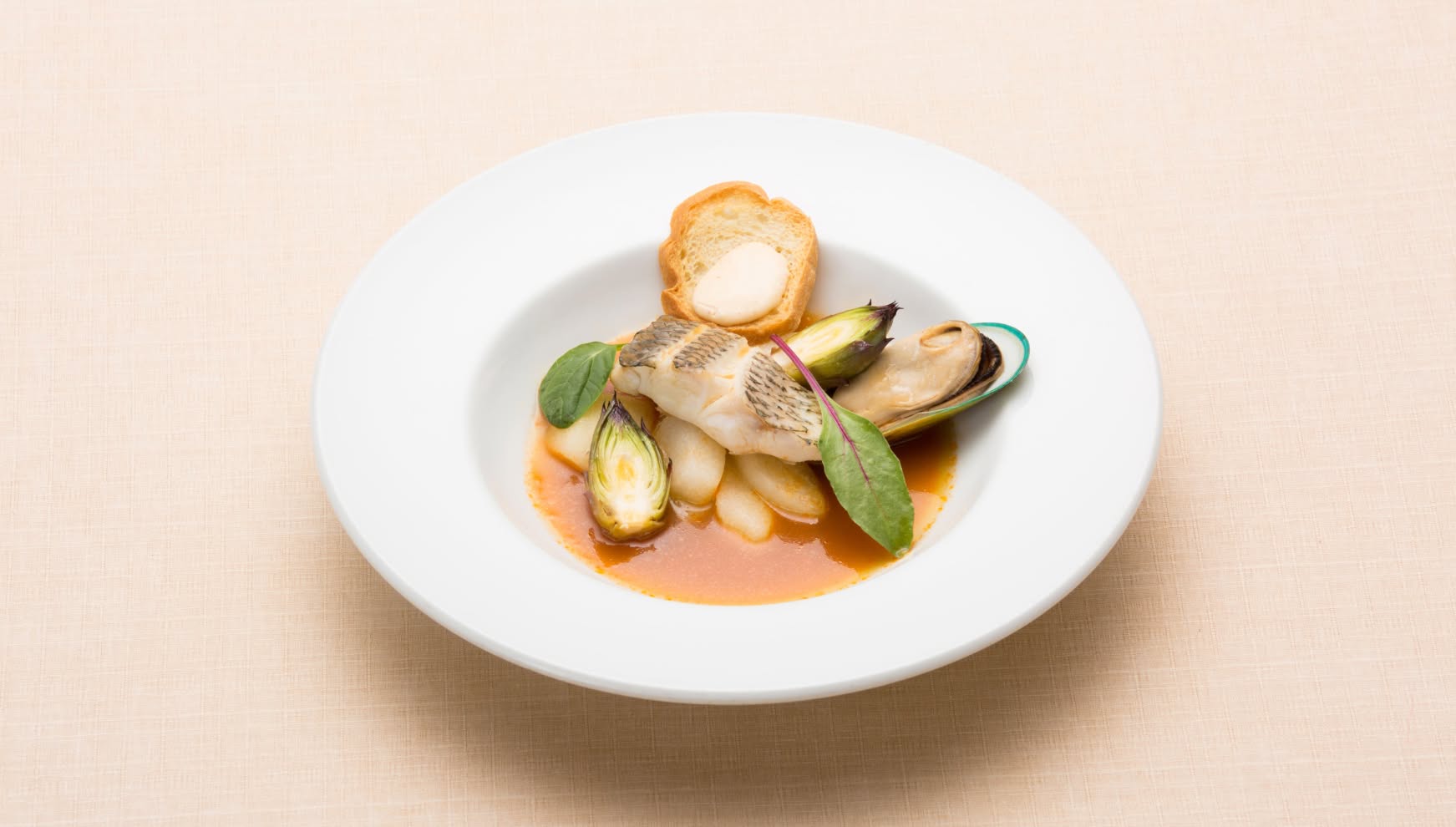 白い深皿に盛り付けられた、アイネス ヴィラノッツェ in カヌチャベイのウエディングメニュー。真鯛のポワレとムール貝に、芽キャベツやハーブが添えられ、旨味が凝縮されたスープとバゲットが調和する、彩り豊かな魚料理の一皿。