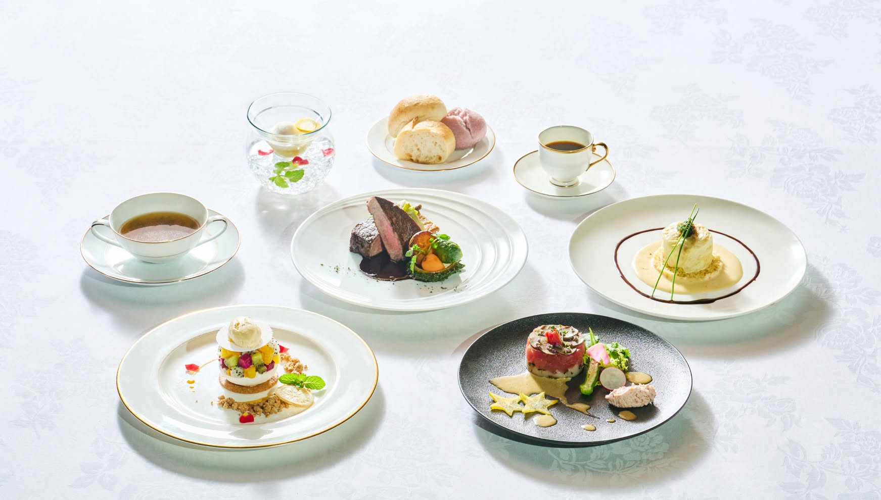 真っ白なテーブルクロスの上に並べられた、ウエディングのフルコース料理一式。前菜のタルタル、魚料理のポワレ、メインの牛フィレ肉、デザートのムース、スープ、パン、コーヒーまでが揃った、華やかで贅沢な祝宴のメニュー。