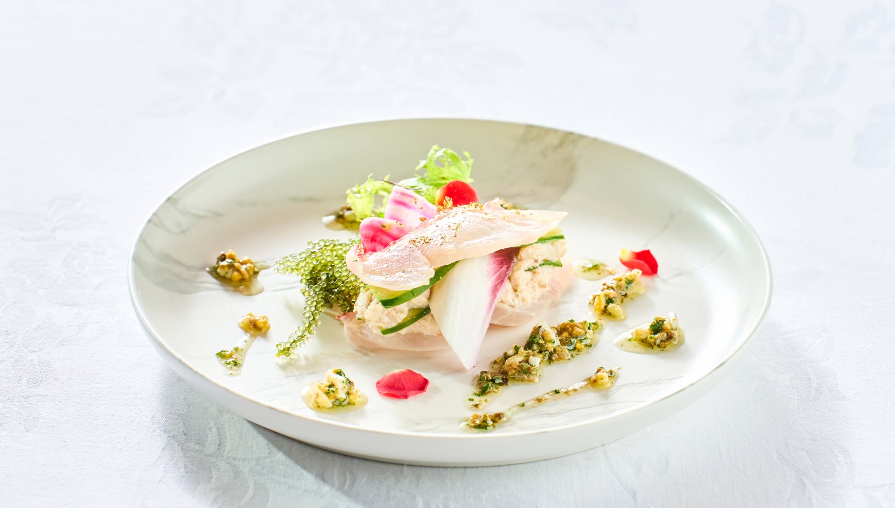 大理石調の白い丸皿に盛り付けられた、海老のムースを鮮魚の切り身で挟んだ華やかな冷製前菜。海葡萄、紅芯大根、エディブルフラワーが彩りを添え、バジルソースで仕上げた、ウエディングの祝宴にふさわしい上品な一皿。