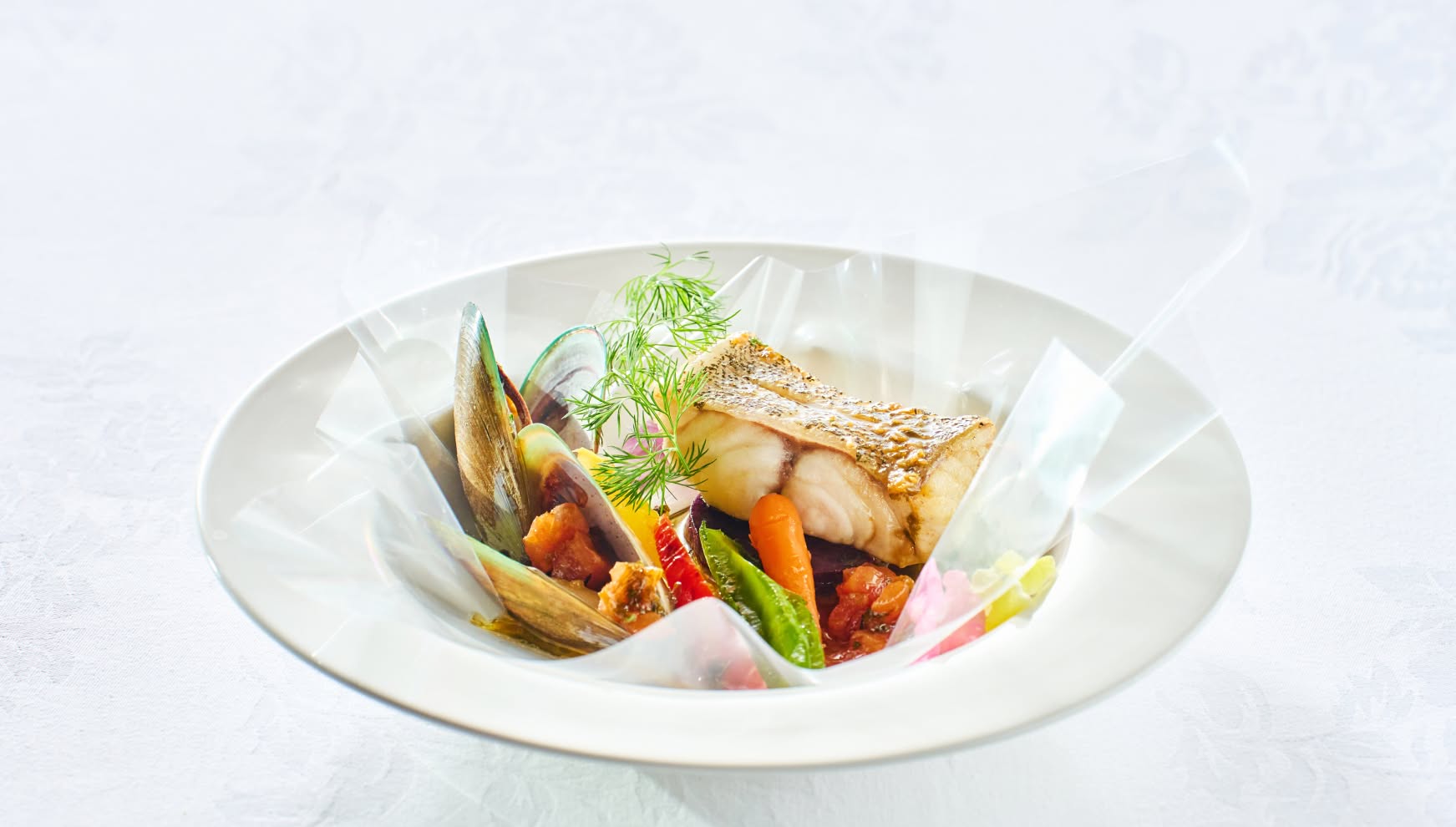 白い深皿に美しく盛り付けられた、鮮魚のポワレとムール貝のアクアパッツァ風料理。耐熱フィルムに包まれた状態で提供され、ディルやミニキャロット、パプリカが彩りを添える、香りと旨味を閉じ込めた華やかなウエディングの魚料理。