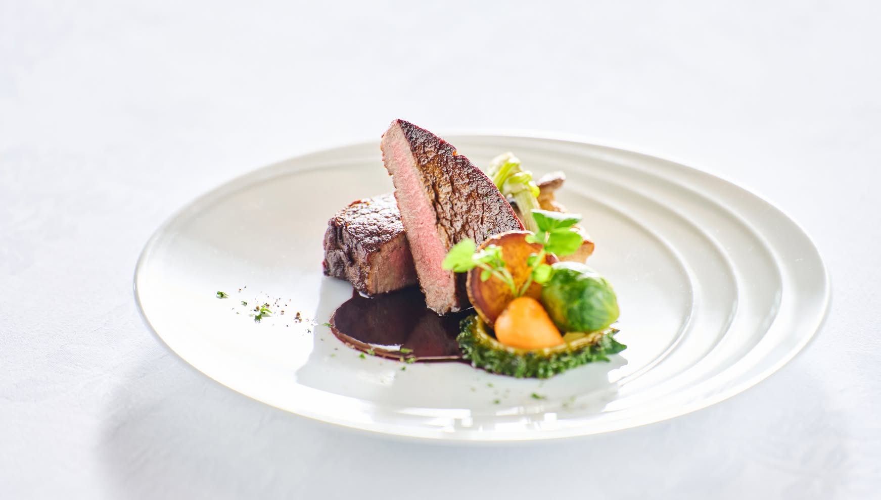 白い波紋のようなデザインの円皿に盛り付けられた、厚切りの牛フィレ肉のロースト。絶妙なミディアムレアの焼き上がりで、濃厚な赤ワインソース、芽キャベツや根菜のローストが添えられた、ウエディングのメインを飾る逸品。