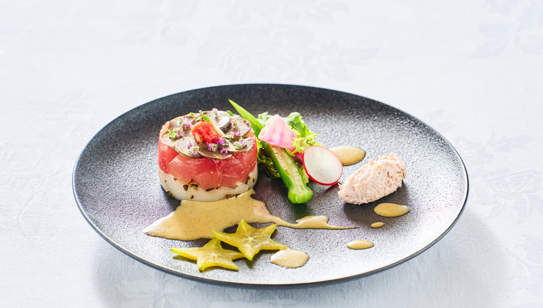 シックな黒の円皿に美しく盛り付けられた、マグロと帆立のタルタル仕立て。トリュフの薄切りやエディブルフラワーを飾り、スターフルーツや新鮮な野菜、ムースを添えた、ウエディングの祝宴を彩る華やかな冷製前菜。