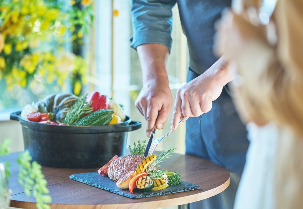 ウエディングの披露宴でシェフが牛肉のステーキをカットする様子。黒いスレート皿にはグリルされた色鮮やかな夏野菜が添えられ、背景には新鮮な野菜が入ったココット鍋が置かれた、素材にこだわる料理の演出。