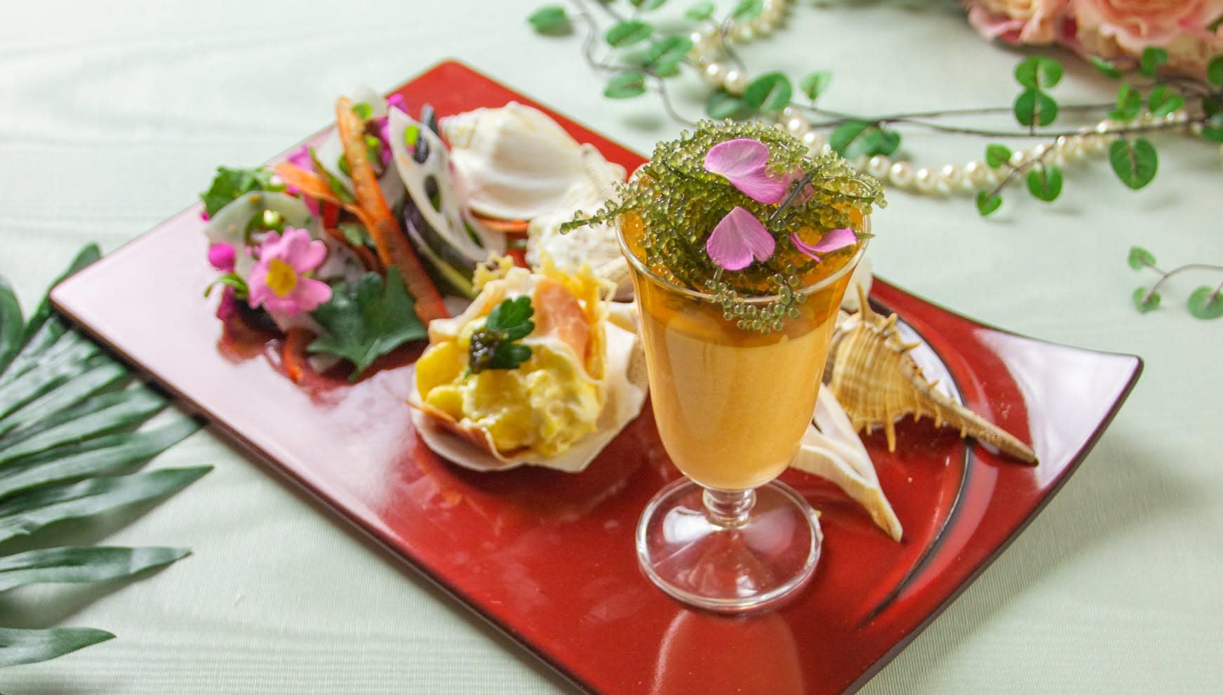 赤を基調とした和モダンな皿に美しく並べられた、ウエディングの祝宴を彩る前菜盛り合わせ。グラスに入った海葡萄を添えたムースを中心に、貝殻に盛り付けられた料理や色鮮やかな食用花が添えられ、パールの装飾が華やかさを演出。