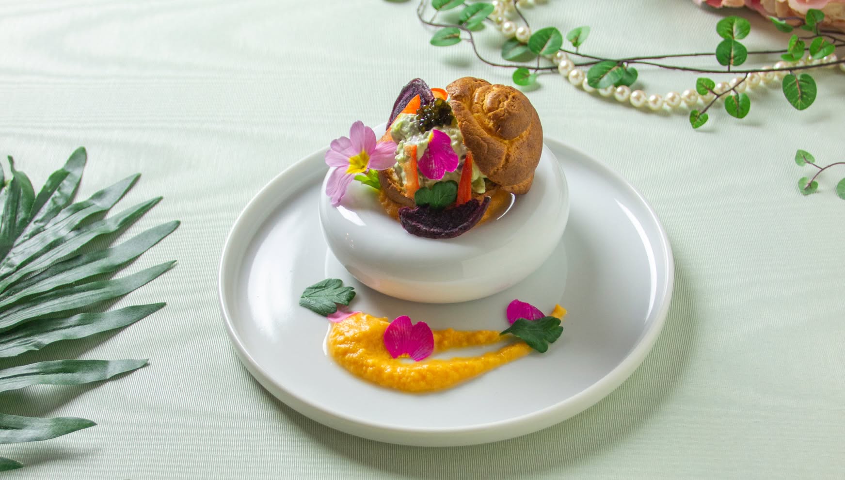 白い丸皿に置かれた小さな白い器に盛り付けられた、華やかな前菜。一口サイズのパンの器にキャビアやエディブルフラワーが飾られ、鮮やかなオレンジ色のソースと花びらが皿を彩る、ウエディングのコース料理を構成する一品。