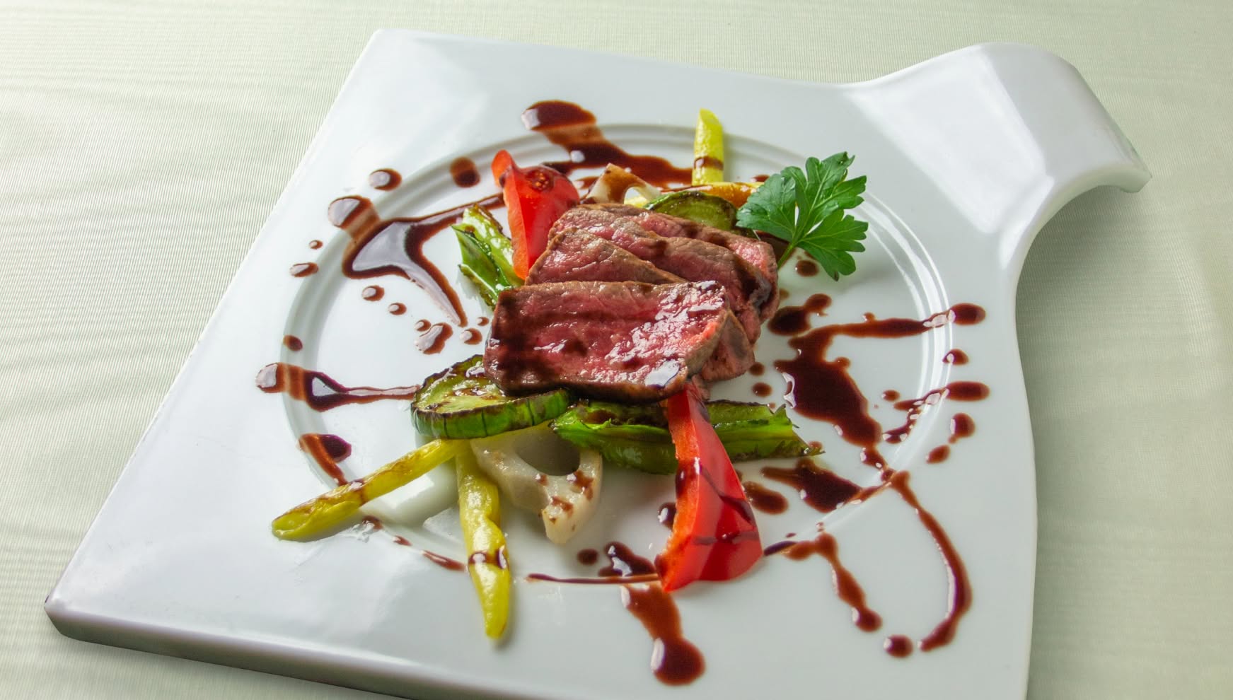 流麗なラインが特徴的な白い変形皿に盛り付けられた、絶妙な火入れの牛フィレ肉のロースト。ズッキーニや赤パプリカなどの焼き野菜が添えられ、バルサミコベースのソースがアーティスティックに描かれた、華やかなウエディングの肉料理。