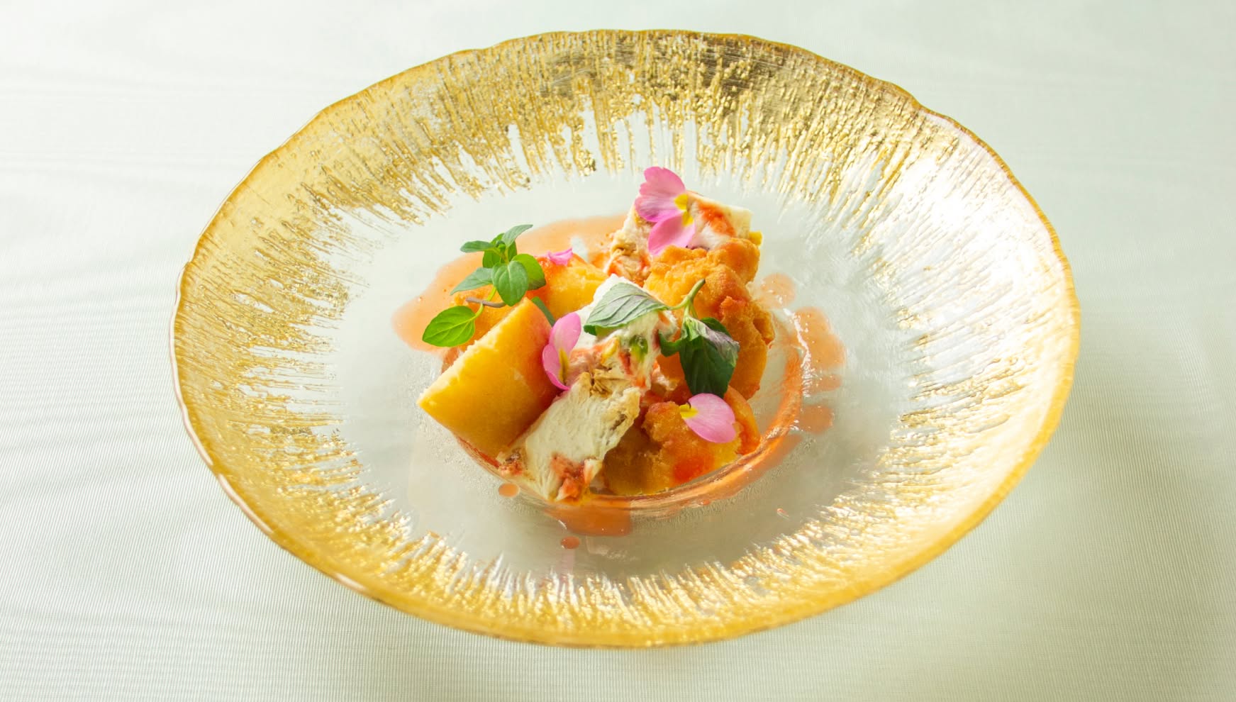 縁がゴールドに輝く華やかなガラス皿に盛り付けられた、ウエディングの最後を飾るデザート。ふんわりとしたムースやフルーツにエディブルフラワーとミントを添え、淡いピンク色のソースで仕上げた、上品で透明感のある一皿。