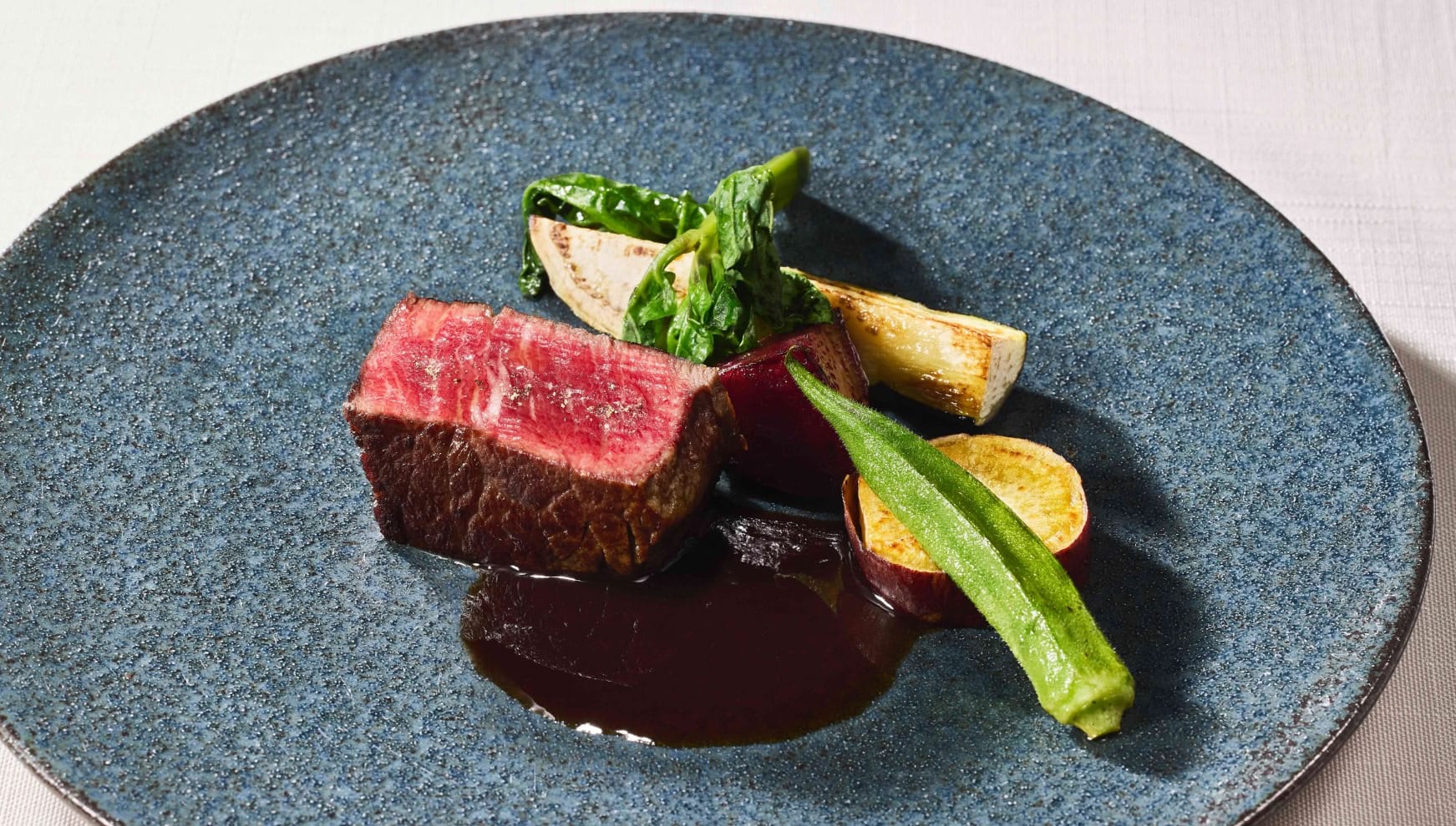 ザ・サーフビーチサイドテラスで提供される、黒毛和牛のグリルをメインとした婚礼料理。絶妙な火入れで鮮やかな赤身を残した厚切り肉に、オクラやサツマイモ、ビーツなどの島野菜が添えられ、深いコクのあるソースで仕上げられた洗練された一皿。