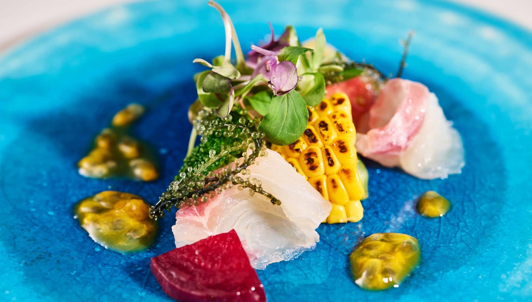 鮮やかなブルーの琉球ガラスのようなプレートに盛り付けられた、沖縄近海魚のカルパッチョ。海ぶどうや焼きトウモロコシ、エディブルフラワーが添えられ、パッションフルーツのソースが彩る。南国らしい素材と色彩が融合した洗練された一皿。