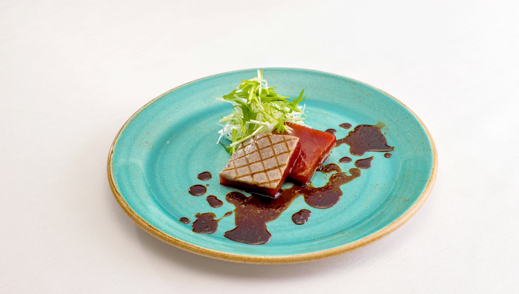 鮮やかなターコイズブルーの平皿に美しく盛り付けられた、表面を香ばしく焼き上げた鮮魚のポワレ。格子状の焼き目が特徴的で、コクのある和風ソースと彩り豊かな水菜のサラダが添えられた、ウエディングコースの魚料理。