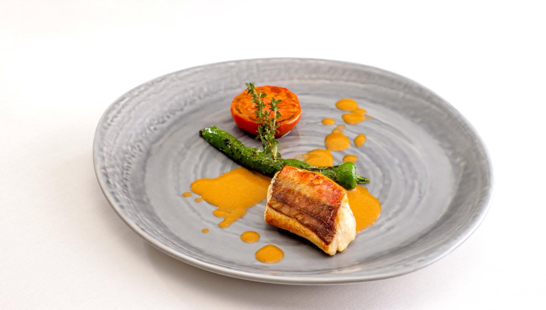 グレーの丸皿に美しく盛り付けられた、皮目をパリッと焼き上げた鮮魚のポワレ。グリルしたトマトとししとう、ハーブを添え、鮮やかなオレンジ色のソースをあしらった、ウエディングコースを彩る上品な魚料理。