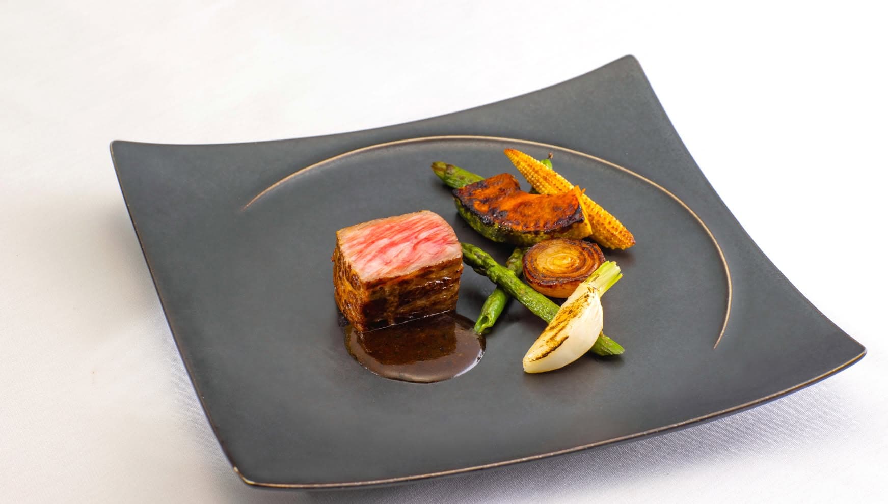 シックな黒の角皿に盛り付けられた、断面が美しいミディアムレアの牛フィレ肉。グリルしたアスパラガス、ヤングコーン、カボチャ、玉ねぎが彩り豊かに添えられ、濃厚なソースが滴るウエディングのメインディッシュ。