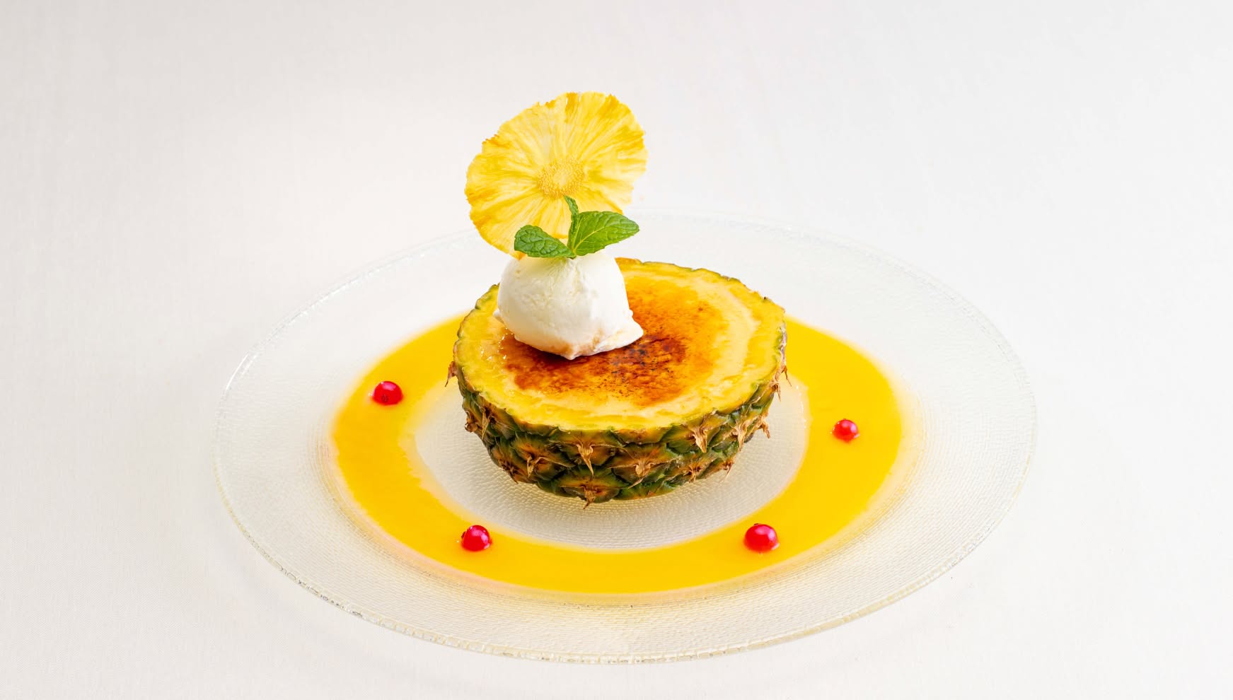 透明なガラス皿に盛り付けられた、パイナップルを器にした華やかなデザート。表面をキャラメリゼしたブリュレにバニラアイスとミント、ドライパイナップルを添え、鮮やかなイエローのソースで彩ったウエディングのスイーツ。