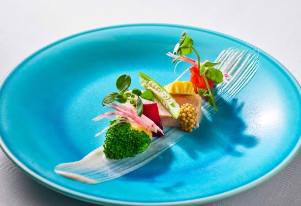 鮮やかなターコイズブルーのプレートに美しく盛り付けられた、婚礼料理の前菜。テリーヌを中心に、ブロッコリーやヤングコーン、紅芋などの色豊かな野菜が添えられ、白いソースのラインが芸術的な一皿。沖縄の海を彷彿とさせる洗練された一品。