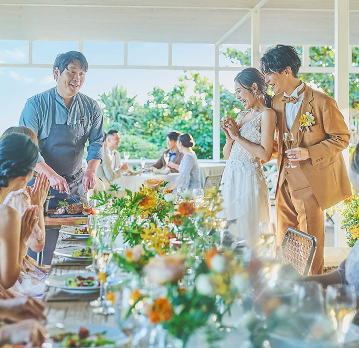 開放的なオーシャンビューの披露宴会場で、シェフが新郎新婦の目の前で肉料理を切り分けるカービングサービス。笑顔で見守る新郎新婦と、華やかに装飾されたテーブル席で食事を楽しむゲスト。ライブ感あふれる演出で、婚礼料理への期待が高まるワンシーン。