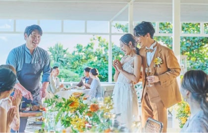 自然光が差し込む披露宴会場で、シェフが新郎新婦の目の前で肉料理を切り分けながら説明し、周囲のゲストが笑顔で拍手している様子