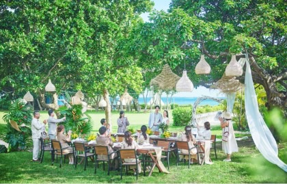 海が見える緑豊かなガーデンでのウエディングパーティー。木から吊るされたラタンの照明の下、新郎新婦が挨拶をし、参列者がテーブルを囲んで笑顔で拍手している和やかな様子