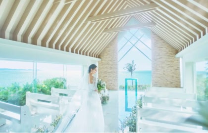 全面ガラス張りで青い海と空が一望できる、光に包まれた明るい挙式会場にて、ブーケを持って静かに佇むウエディングドレス姿の花嫁の後ろ姿。