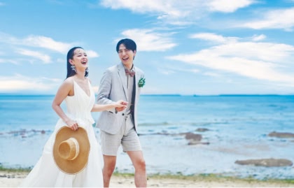 青空の下、海辺で手をつないで楽しそうに大笑いしながら歩く新郎新婦のウエディングフォト。花嫁は麦わら帽子を手に持ち、新郎はショートパンツのカジュアルなスタイル。