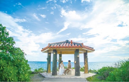 沖縄の青い海と空を背景に、赤瓦の屋根が特徴的な東屋の下で、向かい合って座り見つめ合う新郎新婦