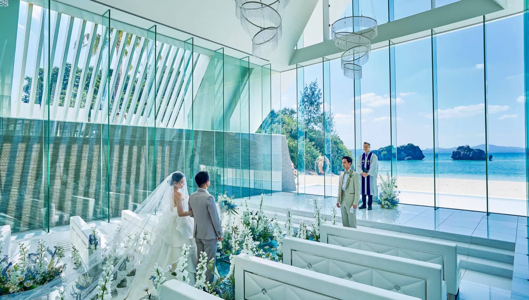 アイネス ヴィラ ノッツェ 沖縄の透明感溢れるチャペルにて、祭壇へ向かう新郎新婦。全面ガラス張りの向こうに広がる青い海と、バージンロードを彩る白い花々、そして二人を待つ牧師。光が差し込む神聖なウエディングの瞬間。