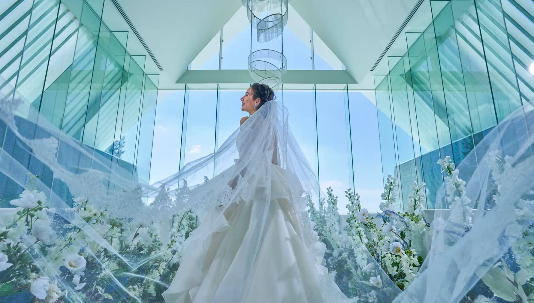 アイネス ヴィラ ノッツェ 沖縄のチャペル。ガラス張りの空間で、新婦のロングベールが風になびき、幻想的に舞い上がる。足元は白い花々に囲まれ、背景の青空と光がウエディングドレスを際立たせる、神秘的な一枚。
