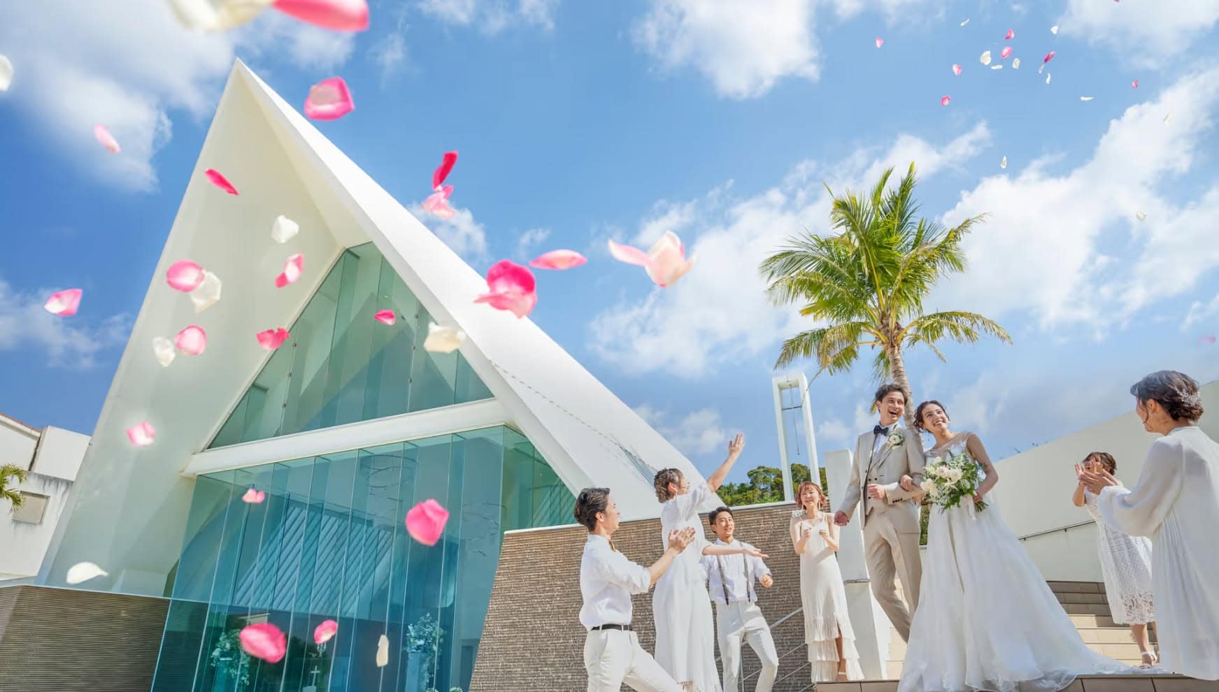 アイネス ヴィラ ノッツェ 沖縄のチャペル外階段にて、ゲストに祝福される新郎新婦。青い空とヤシの木を背に、舞い上がる鮮やかなピンクの花びらの中を笑顔で進む、タキシード姿の新郎とウエディングドレス姿の新婦。