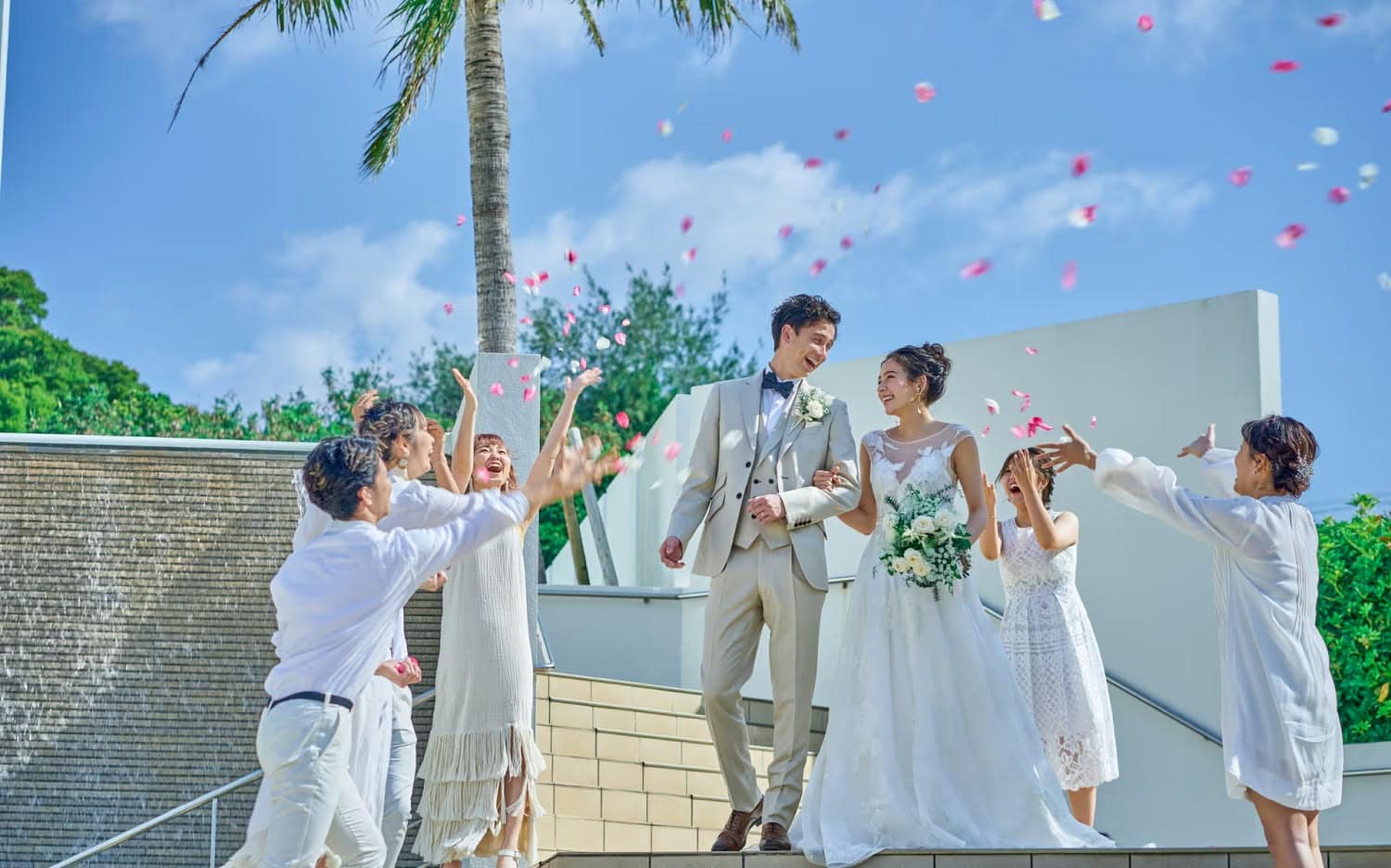 アイネス ヴィラ ノッツェ 沖縄の屋外階段にて、ゲストからフラワーシャワーの祝福を受ける新郎新婦。青空とヤシの木を背景に、笑顔で歩くタキシード姿の新郎とウエディングドレス姿の新婦、それを見守る参列者の幸せな光景。