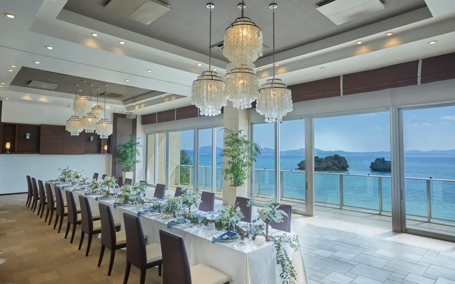 アイネス ヴィラ ノッツェ 沖縄のパーティ会場。大きな窓から沖縄の青い海を一望できる開放的な空間に、白を基調とした装花が施されたロングテーブルが配置されている。天井には華やかなシャンデリアが輝く上質な空間。