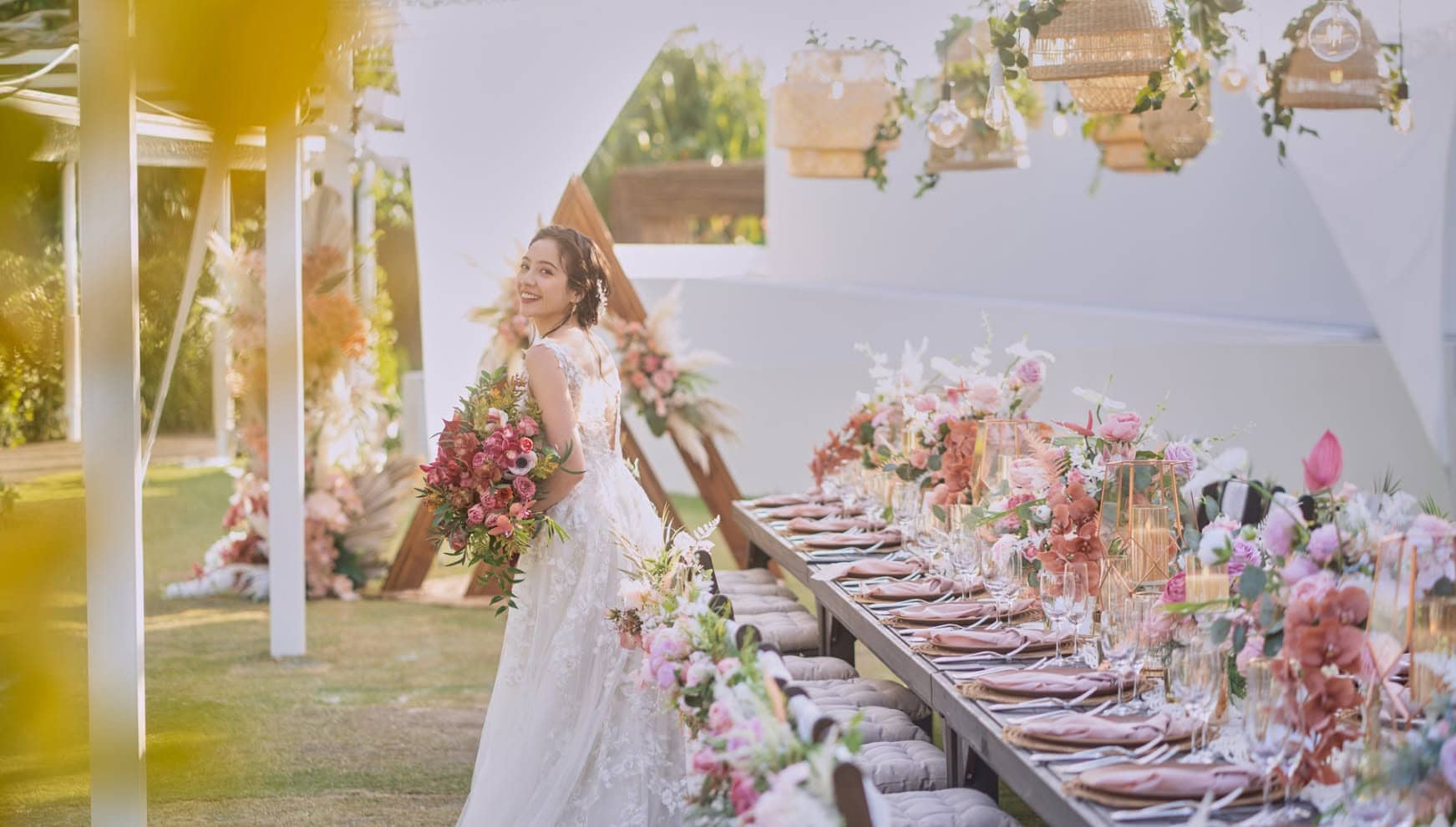 沖縄の結婚式場「アイネス ヴィラノッツェ 沖縄」のガーデンにて、華やかな装飾に囲まれた披露宴会場。手前にはピンクや白の花々で彩られた長いテーブルセットが並び、色鮮やかなブーケを手にした花嫁が微笑みながら振り返っています。温かみのあるカゴ状のペンダントライトが吊るされた、開放的でナチュラルな空間が広がっています。