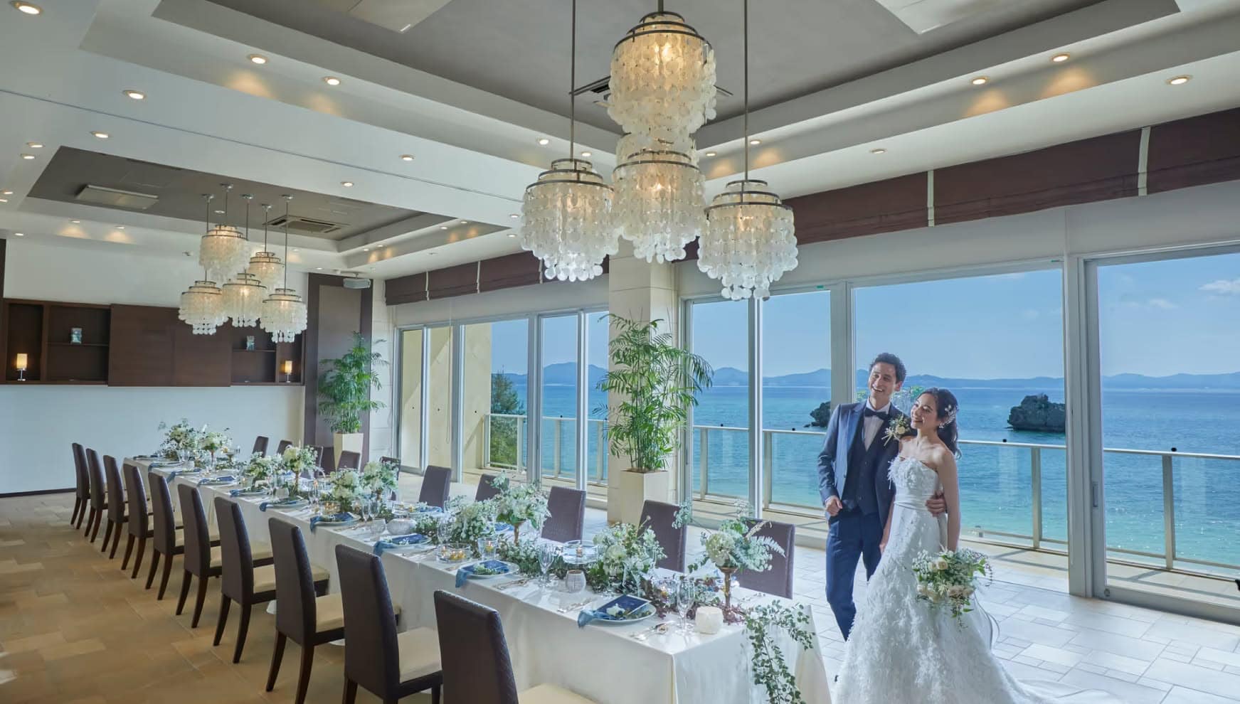 アイネス ヴィラ ノッツェ 沖縄のパーティ会場にて、オーシャンビューを背に笑顔を見せる新郎新婦。華やかなシャンデリアが輝く会場内には、海の色に合わせたブルーのナプキンと白の花々で彩られた美しいテーブルセットが広がる。